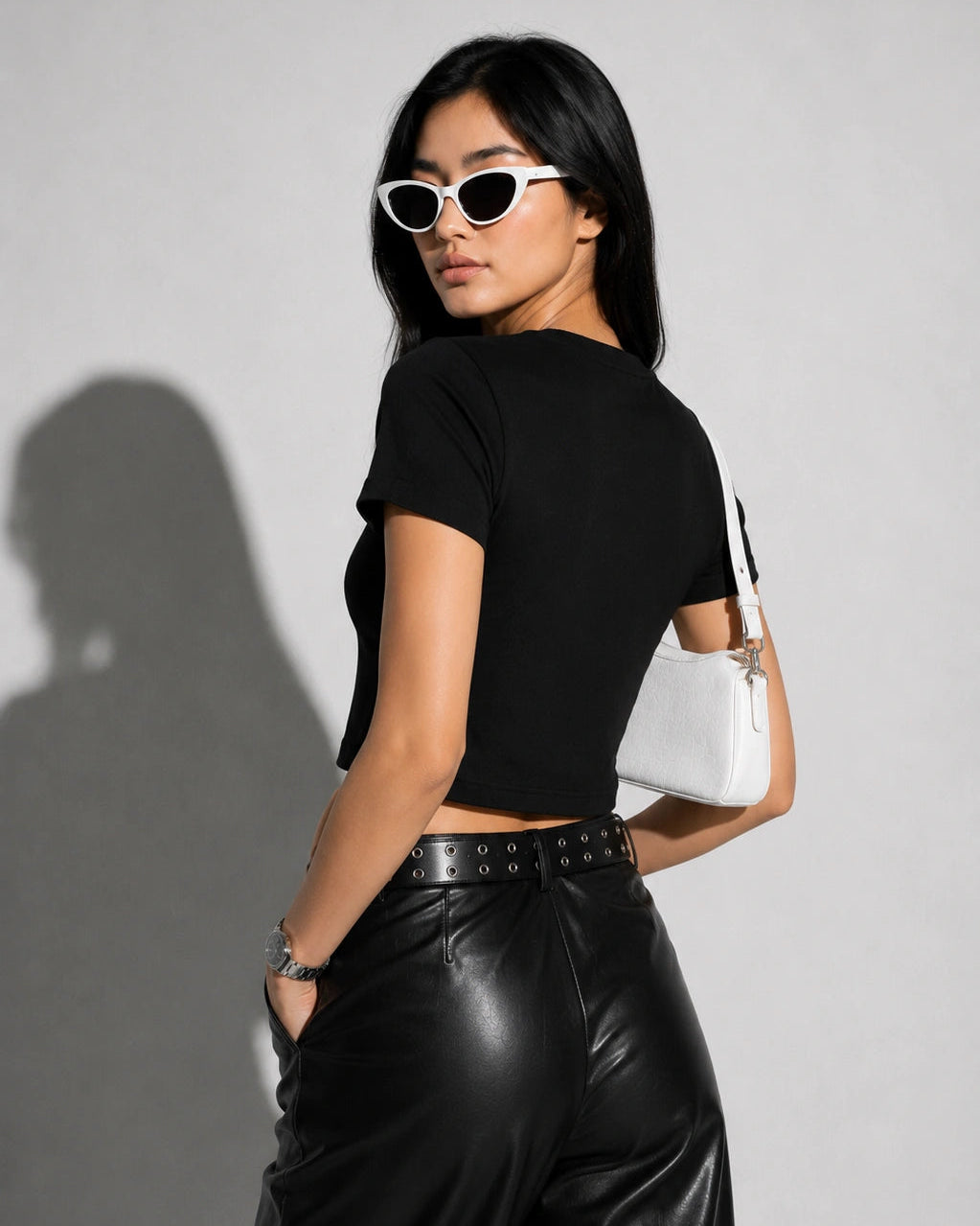 Woman wearing a black crop top and leather pants with a white handbag against a gray background