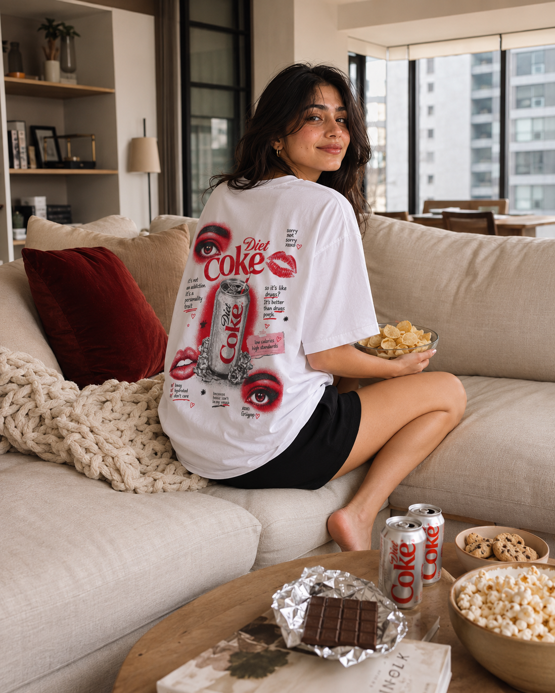 Person sitting on a couch wearing a white t-shirt with a Diet Coke design, holding popcorn and a can of Diet Coke.