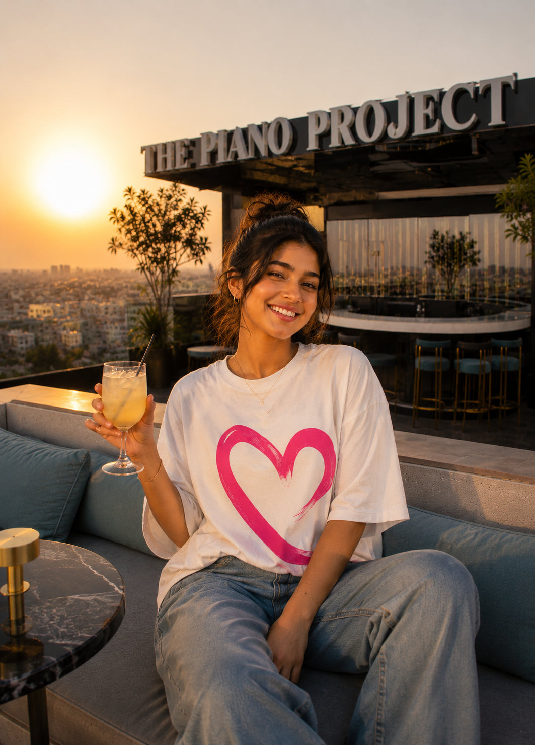 Woman sitting on a rooftop bar with a drink, wearing a white t-shirt with a pink heart design, at sunset.