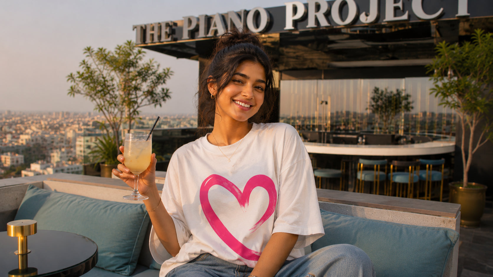Woman holding a drink with a heart design on her shirt at a rooftop bar with 'The Piano Project' sign.